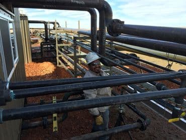 Worker in hard hat inspecting industrial piping system against blue sky background.