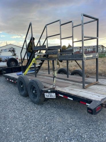 Metal frame structure on a trailer, secured with straps, against a rural backdrop.