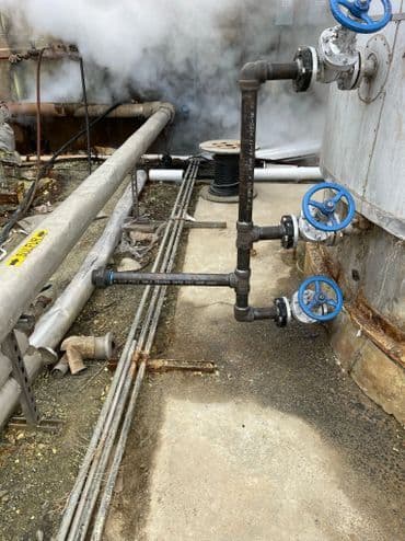 Industrial pipes and valves with blue handles amidst steam on a factory floor.