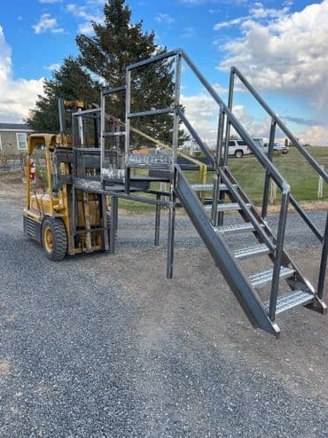 Forklift positioned near metal loading ramp stairs on gravel site with scenic background.