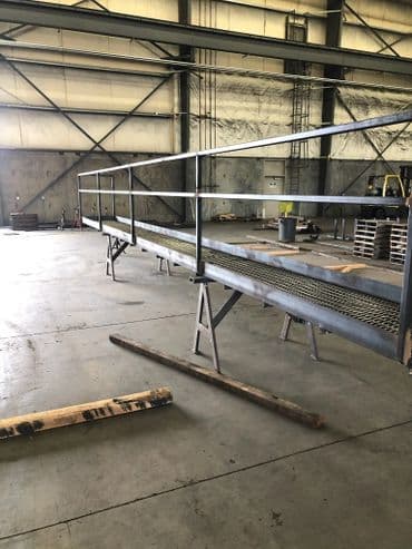 Steel walkway under construction in a warehouse with wooden supports and pallets.