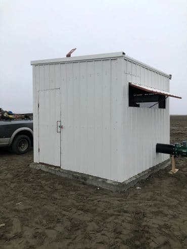 White metal storage shed on a sandy ground with a window and door for access.
