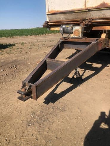 Metal trailer hitch with a sturdy frame, located on a dirt field with green crops in the background.