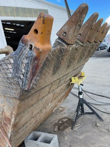 Excavator bucket with wear and tear on cutting edges, positioned on a worksite.