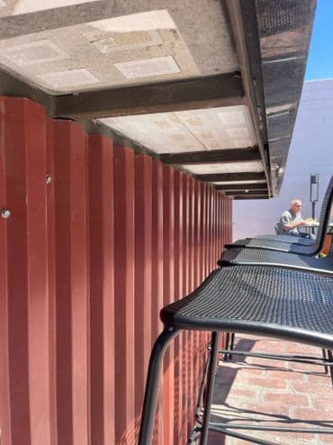 Bar stools lined up against a red metal wall, with a person seated in the background.