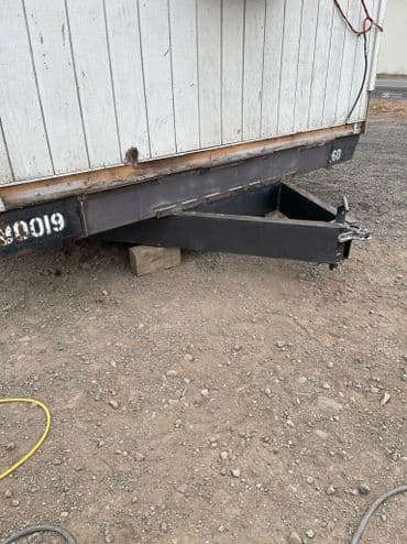 Trailer hitch mounted on a metal frame, resting on a gravel surface with exposed wiring.