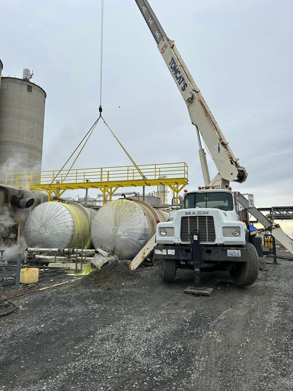 Mack truck lifting industrial tanks at a construction site with crane equipment.