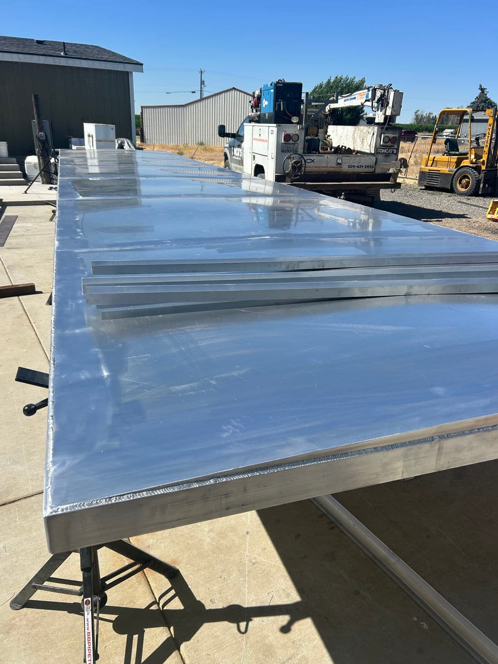 Aluminum sheet being prepared on a work table with machinery and buildings in the background.