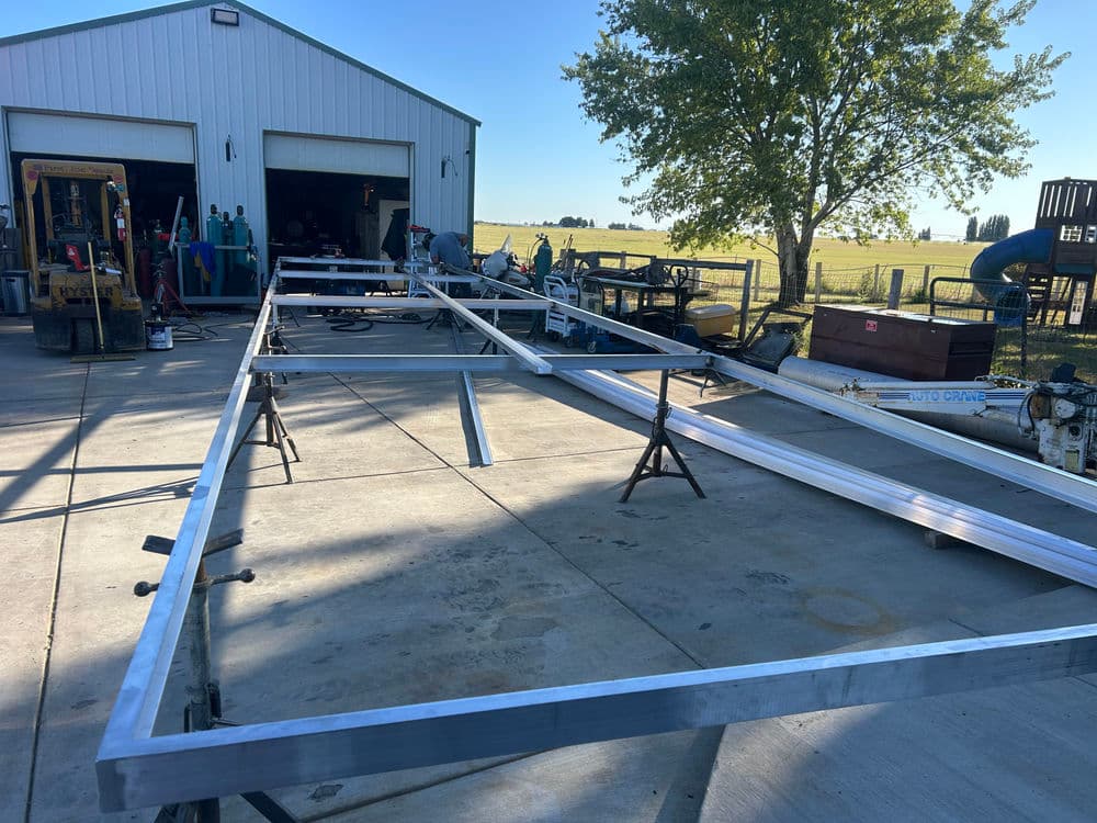 Aluminum frame structure under construction in a workshop with machinery and grass field background.