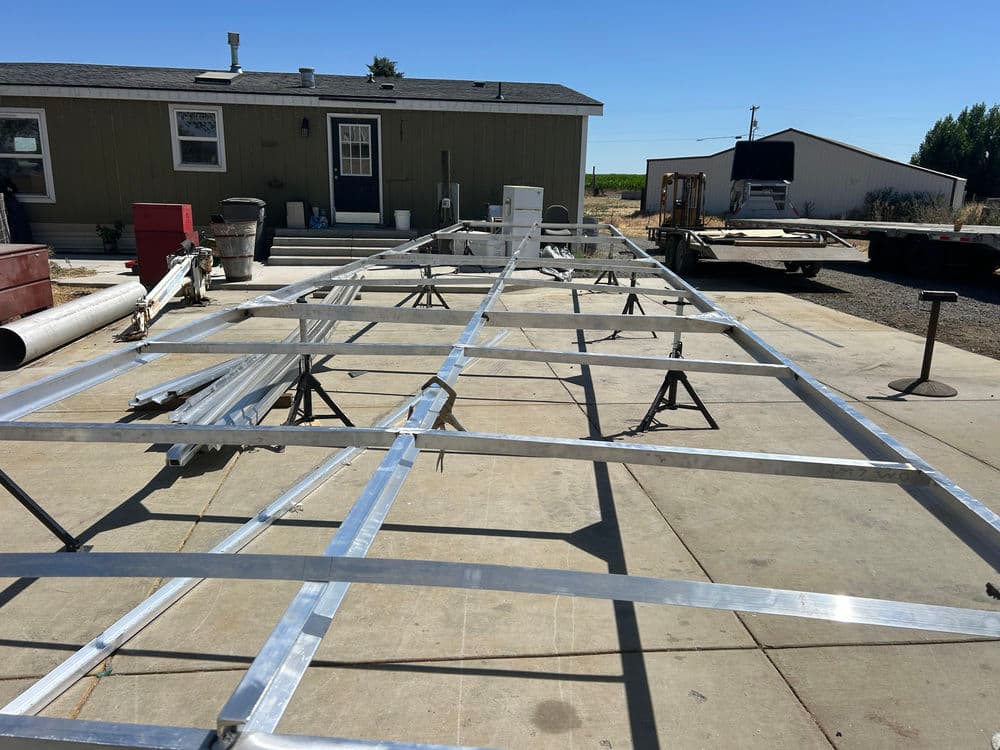 Aluminum trailer frame under construction on a sunny day with workshop in background.