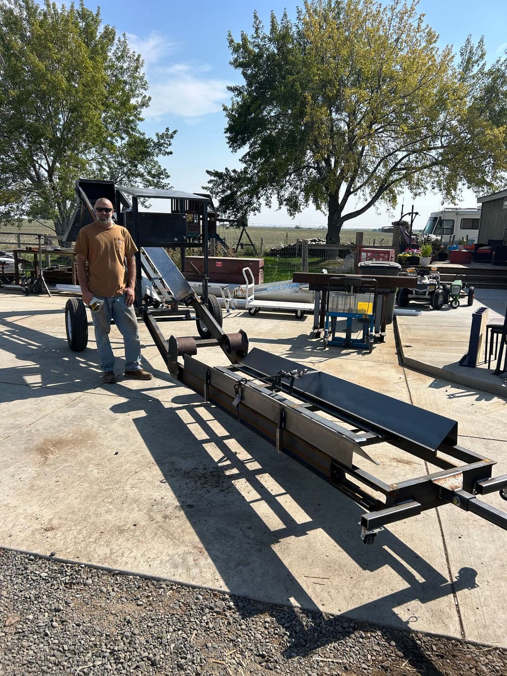 Man operating a metal conveyor on a farm, surrounded by agricultural equipment and trees.