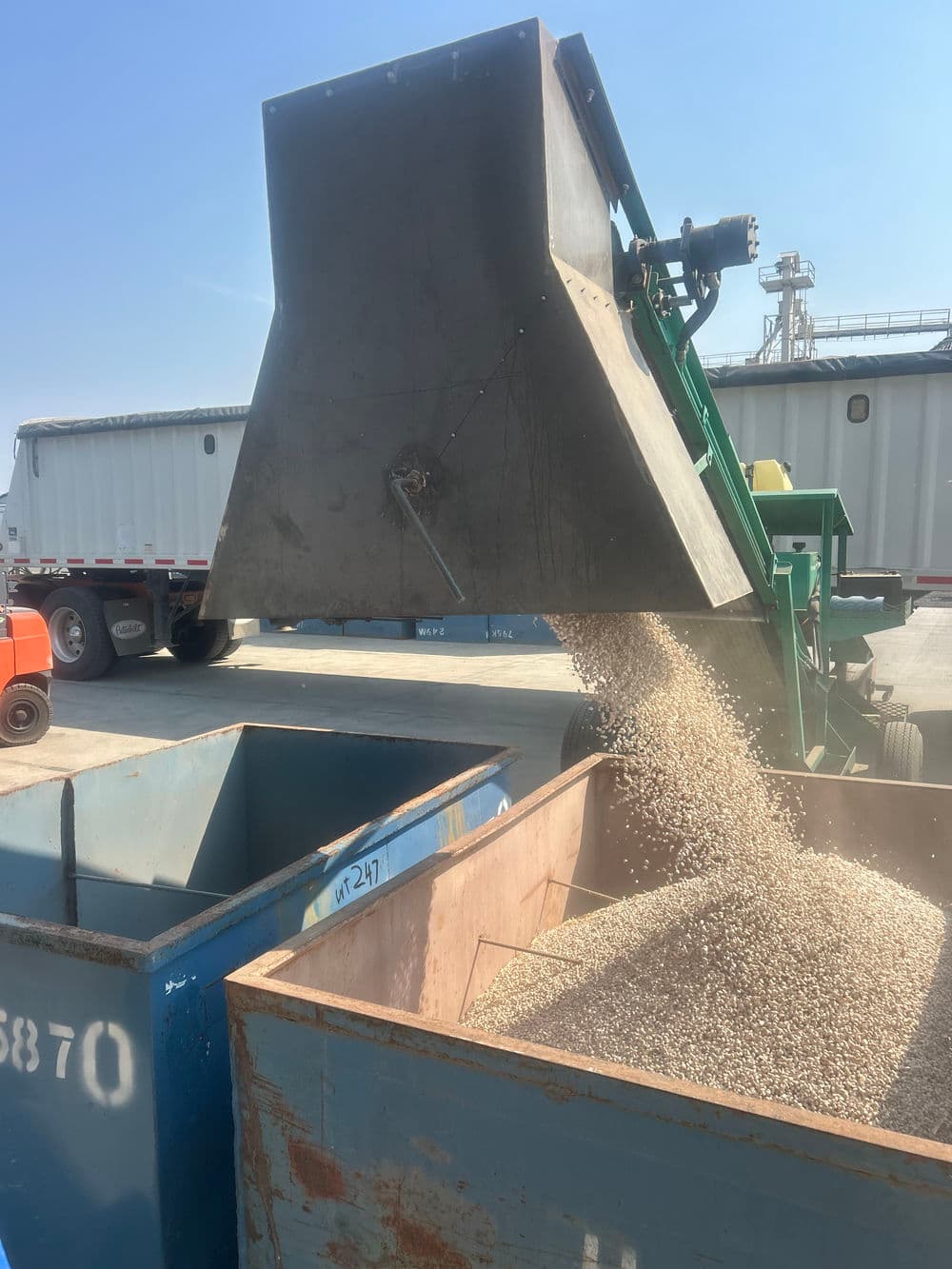 Machine pouring grain into blue containers at a loading facility on a sunny day.